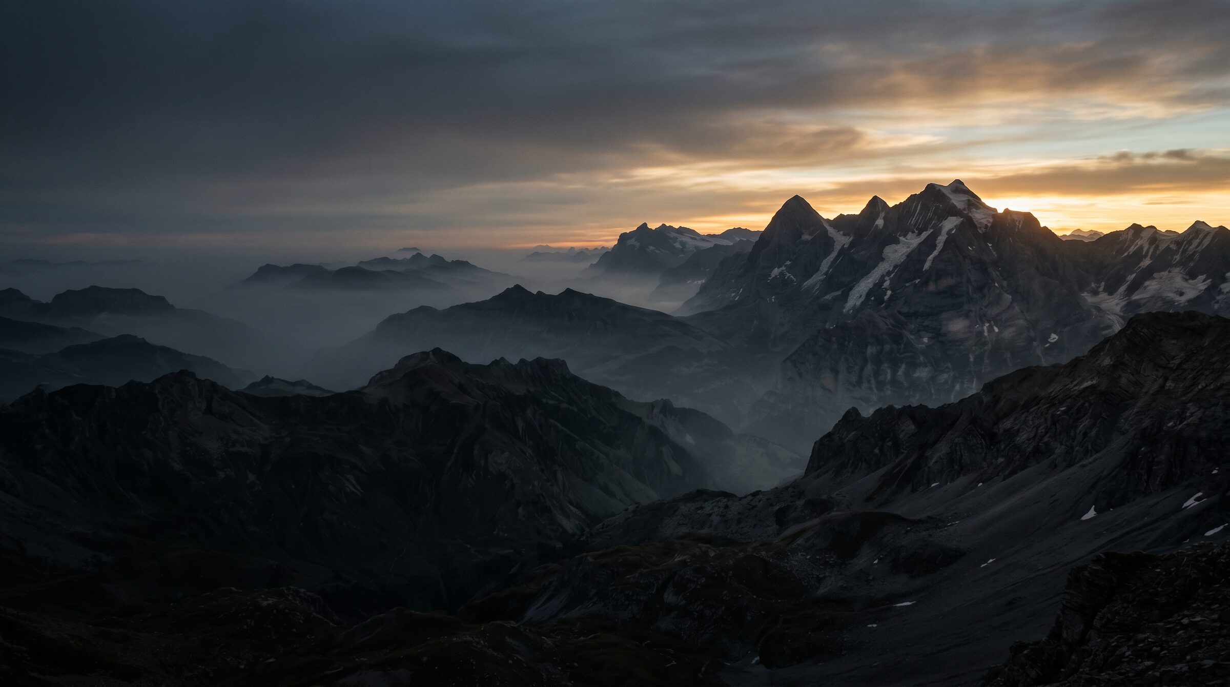 A dark mountain range at dawn, mist settling in the valleys below a distant lit summit